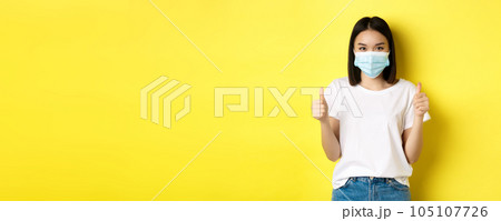 Covid-19, quarantine and social distancing concept. Cheerful asian woman in medical mask and white t-shirt showing thumbs up in approval, praise good deal, yellow background 105107726