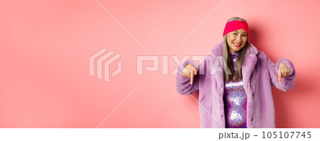 Fashion and shopping concept. Smiling senior asian woman pointing fingers down, showing store advertisement on bottom, wearing stylish purple winter coat, pink background Fashion and shopping concept. Smiling senior asian woman pointing fingers down, showing store advertisement on bottom, wearing stylish purple winter coat, pink background 105107745
