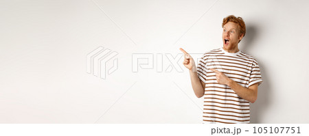 Check it out. Amazed redhead guy pointing and looking left at promotion banner, standing over white background 105107751