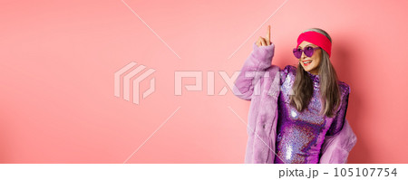 Asian old woman in fashionable glittering dress and purple faux fur coat dancing disco, pointing finger up and having fun, pink background Asian old woman in fashionable glittering dress and purple faux fur coat dancing disco, pointing finger up and having fun, pink background 105107754