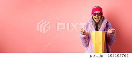 Shopping and fashion concept. Stylish asian elderly woman in sunglasses and faux fur coat open paper bag with gifts, looking amazed at camera, pink background Shopping and fashion concept. Stylish asian elderly woman in sunglasses and faux fur coat open paper bag with gifts, looking amazed at camera, pink background 105107763