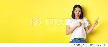 Beauty and fashion concept. Beautiful asian woman in white t-shirt pointing fingers right, demonstrate logo standing over yellow background Beauty and fashion concept. Beautiful asian woman in white t-shirt pointing fingers right, demonstrate logo standing over yellow background 105107764