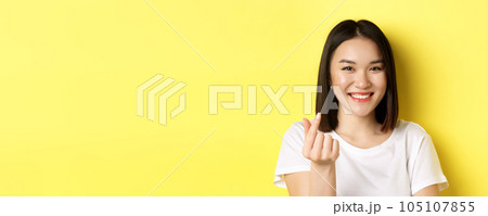 Valentines day and women concept. Close up of pretty asian girl in white t-shirt, smiling and showing finger heart, standing over yellow background Valentines day and women concept. Close up of pretty asian girl in white t-shirt, smiling and showing finger heart, standing over yellow background 105107855