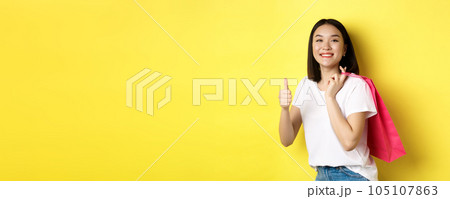 Cheerful asian female shopper looking amused, holding shopping bag and showing thumbs up, recommend store with discounts, standing over yellow background 105107863