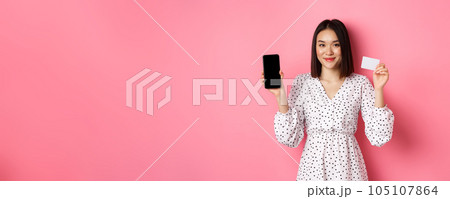 Cute asian woman shopping online, showing bank credit card and mobile screen, smiling and looking at camera, standing over pink background Cute asian woman shopping online, showing bank credit card and mobile screen, smiling and looking at camera, standing over pink background 105107864
