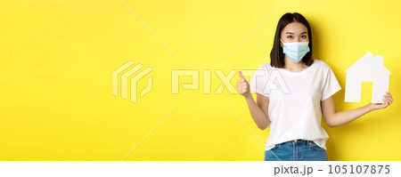 Covid-19, pandemic and real estate concept. Pretty asian woman smiling in medical mask, showing paper house cutout and thumb up, approve and like agency for buying property 105107875