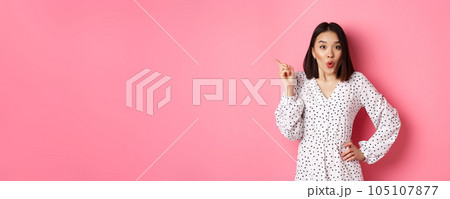 Excited asian female model showing promo offer, pointing at upper left corner and staring at camera amazed, wearing trendy spring dress, standing over pink background 105107877