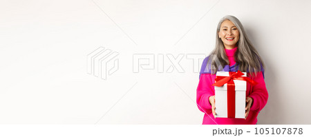 Beautiful asian senior woman smiling, congratulating with valentines day, holding gift in box, standing over white background Beautiful asian senior woman smiling, congratulating with valentines day, holding gift in box, standing over white background 105107878