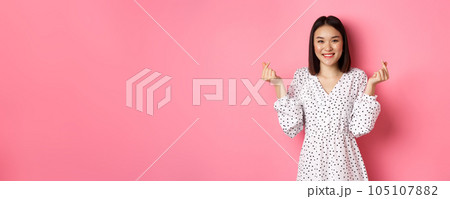 Lovely asian woman in dress showing korean heart signs and smiling, standing on romantic pink background Lovely asian woman in dress showing korean heart signs and smiling, standing on romantic pink background 105107882