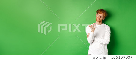 Young man with red messy hair and glasses, smiling pleased and pointing hand at camera, your turn gesture, standing over green background Young man with red messy hair and glasses, smiling pleased and pointing hand at camera, your turn gesture, standing over green background 105107907