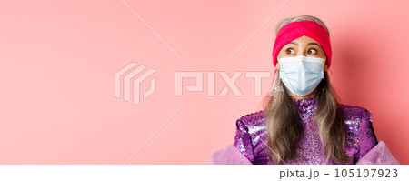 Covid-19, virus and fashion concept. Surprised and amazed asian grandmother in stylish outfit and medical mask, looking left impressed, pink background 105107923