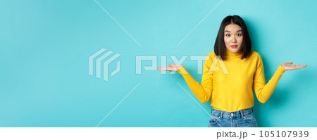 Image of indecisive asian woman shrugging shoulders, spread hands sideways and looking clueless at camera, standing indecisive against blue background 105107939