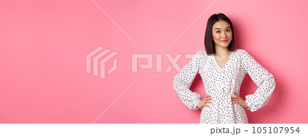 Beautiful korean lady in dress looking at camera and smiling, waiting for something, standing against pink background Beautiful korean lady in dress looking at camera and smiling, waiting for something, standing against pink background 105107954