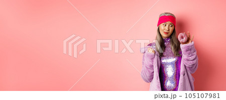 Funny asian senior woman in glittering disco dress and faux fur coat looking tempted at delicious donut, wanting to eat sweet, standing over pink background 105107981