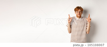 Hopeful young man with red hair and glasses cross fingers for good luck, making wish, standing over white background 105108007