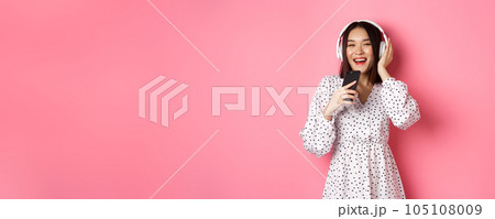 Beautiful smiling asian woman singing song in smartphone microphone, playing karaoke app and using headphones, standing over pink background Beautiful smiling asian woman singing song in smartphone microphone, playing karaoke app and using headphones, standing over pink background 105108009