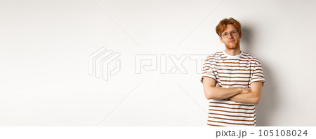 Teenage redhead guy in glasses cross arms on chest, looking skeptical and unamused at camera, standing over white background 105108024