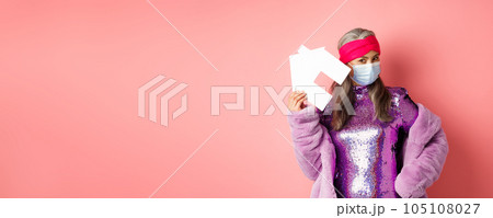 Covid-19 and real estate concept. Cool and stylish asian elderly woman in disco dress and medical mask showing paper house, standing over pink background Covid-19 and real estate concept. Cool and stylish asian elderly woman in disco dress and medical mask showing paper house, standing over pink background 105108027