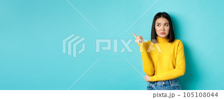 Shopping concept. Disappointed and gloomy asian woman sulking upset, pointing finger left at bad news banner, standing over blue background 105108044
