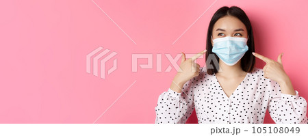 Covid-19, pandemic and lifestyle concept. Kawaii asian girl pointing fingers at her face mask, wearing preventive measures from coronavirus, smiling with eyes, pink background Covid-19, pandemic and lifestyle concept. Kawaii asian girl pointing fingers at her face mask, wearing preventive measures from coronavirus, smiling with eyes, pink background 105108049