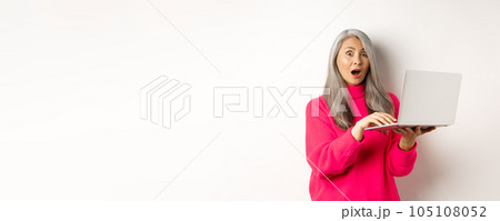 Surprised and impressed asian grandmother staring shocked at camera after reading news on laptop screen, standing over white background Surprised and impressed asian grandmother staring shocked at camera after reading news on laptop screen, standing over white background 105108052