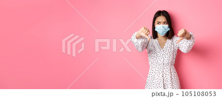 Coronavirus, social distancing and lifestyle concept. Disappointed asian woman in face mask looking judgemental, showing thumbs-down, express negative opinion, pink background Coronavirus, social distancing and lifestyle concept. Disappointed asian woman in face mask looking judgemental, showing thumbs-down, express negative opinion, pink background 105108053