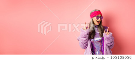 Hipster granny in sunglasses and glittering dress posing for photo with peace, victory signs, standing sassy against pink background 105108056