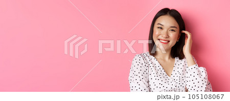 Close-up of beautiful asian woman smiling happy, touching new haircut, standing over pink background 105108067