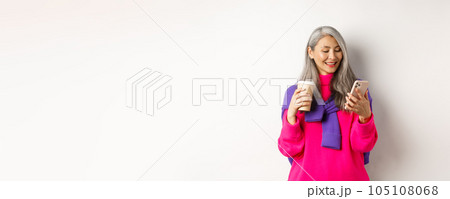 Trendy senior woman drinking coffee and using smartphone. Korean senior lady reading message on mobile phone and smiling, white background 105108068