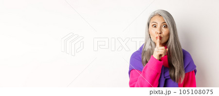Close-up of stylish senior asian woman in hipster outfit telling hush, shushing at looking at camera, show taboo gesture, standing over white background 105108075