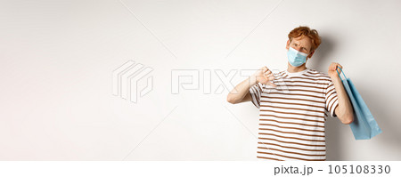 Concept of social distancing and shopping. Disappointed young man with red hair, wearing face mask, holding shopping bag and showing thumb-down from dislike, disapprove store 105108330