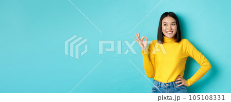 Impressed asian woman in yellow sweater, looking left amazed and showing OK sign in approval, standing over blue background Impressed asian woman in yellow sweater, looking left amazed and showing OK sign in approval, standing over blue background 105108331