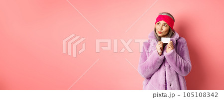 Shopping and fashion concept. Fashionable asian senior woman thinking about buying gifts, holding credit card and looking thoughtful at upper left corner, pink background Shopping and fashion concept. Fashionable asian senior woman thinking about buying gifts, holding credit card and looking thoughtful at upper left corner, pink background 105108342