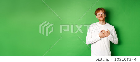 Handsome young man in glasses and white long-sleeve smiling, holding hands on heart with grateful face, thanking you, standing over green background 105108344