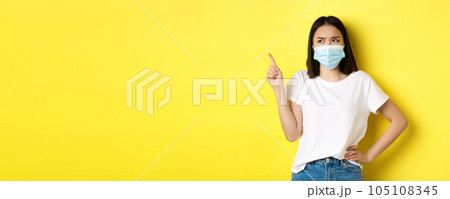 Covid, health care and pandemic concept. Asian woman in medical mask and white t-shirt pointing finger at upper left corner logo, showing promotion, yellow background 105108345