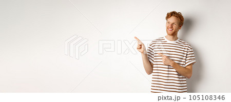 Cheerful caucasian guy with red hair and beard, looking and pointing left at logo banner, standing over white background 105108346