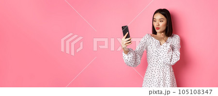 Beautiful asian girl using photo filters app and taking selfie on smartphone, posing in cute dress against pink background 105108347