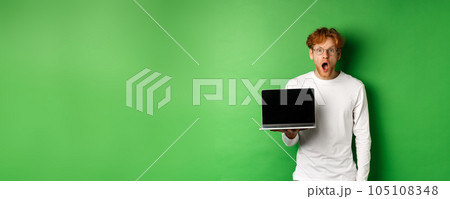 Impressed redhead guy in glasses drop jaw after seeing online promo, showing laptop screen and staring at camera with disbelief, standing over green background 105108348