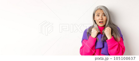 Close up of scared asian senior woman gasping, looking frightened and startled at camera, standing over white background 105108637