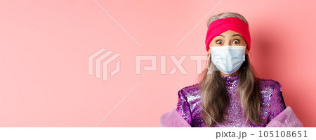 Covid-19, virus and social distancing concept. Close up of fashionable elderly woman in medical mask and headband staring surprised at camera, pink background Covid-19, virus and social distancing concept. Close up of fashionable elderly woman in medical mask and headband staring surprised at camera, pink background 105108651