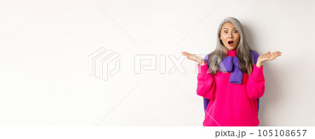 Surprised and amazed asian elderly woman spread hands sideways in complete disbelief, gasping and staring impressed at camera, standing over white background Surprised and amazed asian elderly woman spread hands sideways in complete disbelief, gasping and staring impressed at camera, standing over white background 105108657