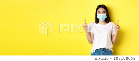 Covid-19, pandemic and social distancing concept. Young asian woman in white t-shirt and medical mask from coronavirus, pointing fingers up, showing special offer 105108658