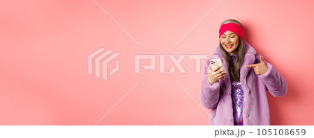 Online shopping and fashion concept. Cool asian senior woman checking out promotion on mobile phone, pointing finger at smartphone screen and smiling pleased, pink background 105108659