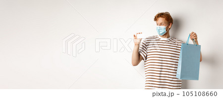 Concept of covid-19 and lifestyle. Cheerful young man with red hair, wear medical mask, showing shopping bag from store and plastic credit card 105108660