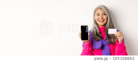 Online shopping. Closeup of smiling asian senior woman showing blank mobile screen and plastic credit card, looking happy, standing over white background Online shopping. Closeup of smiling asian senior woman showing blank mobile screen and plastic credit card, looking happy, standing over white background 105108662