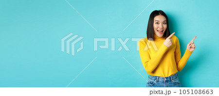 Shopping concept. Beautiful chinese girl in yellow sweater pointing fingers at upper right corner logo banner, smiling amused, standing over blue background Shopping concept. Beautiful chinese girl in yellow sweater pointing fingers at upper right corner logo banner, smiling amused, standing over blue background 105108663