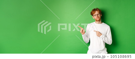 Doubtful redhead guy in glasses pointing fingers left, looking hesitant and unsure at camera, standing over green background Doubtful redhead guy in glasses pointing fingers left, looking hesitant and unsure at camera, standing over green background 105108695