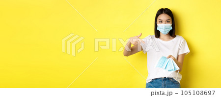 Coronavirus, quarantine and medicine concept. Young asian woman pointing finger at medical masks for going outdoors, standing over yellow background 105108697