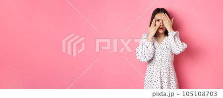 Disappointed asian woman peeking through fingers, open eyes and frowning displeased, staring with disdain at camera, standing over pink background 105108703