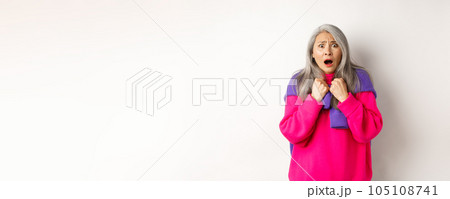 Scared and shocked asian grandmother gasping and staring frustrated at camera, standing against white background Scared and shocked asian grandmother gasping and staring frustrated at camera, standing against white background 105108741